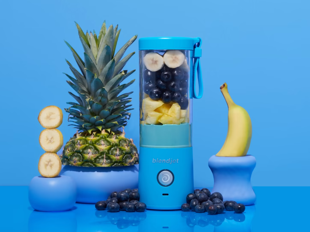 A bright blue BlendJet blender filled with banana slices, blueberries, and pineapple chunks, surrounded by whole fruits including a pineapple, banana, and blueberries, set against a vivid blue background.