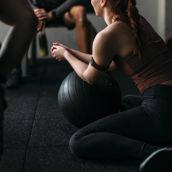  A person wearing a Polar optical heart rate sensor on their upper arm, visible through the red adjustable strap. The individual is seated on the floor in a gym, resting their arms on a large black exercise ball. The person is dressed in athletic wear, with another individual in workout attire visible in the background, emphasizing the fitness context.