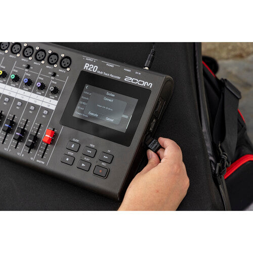 A hand adjusting the knob on the Zoom R20 audio mixer and recorder, which is displayed on a dark surface. The device's digital screen shows the menu options for audio settings, and multiple channels with faders and knobs are visible. This setup demonstrates the user-friendly interface and the hands-on control features of the audio equipment.