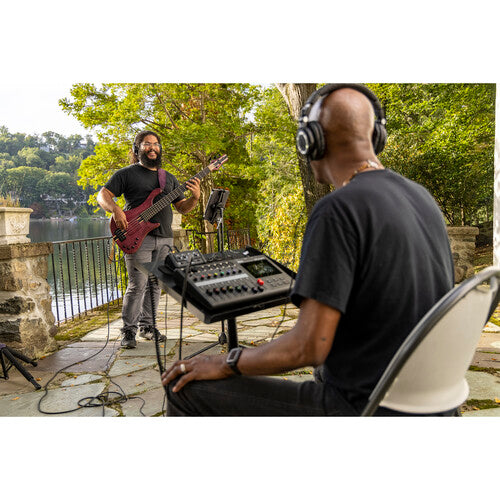 A man playing an electric bass guitar outdoors while another man, wearing headphones and seated on a chair, operates an audio mixer. The setting appears to be a scenic patio overlooking a lake, surrounded by trees and a stone railing. The scene conveys a relaxed outdoor recording session or live performance.