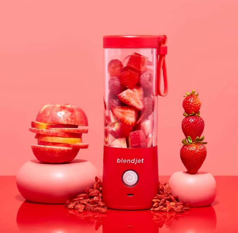 A vibrant red BlendJet blender filled with fresh strawberries, apples, and raspberries, surrounded by stacked apple slices, whole strawberries, and dried goji berries on a bold red background.