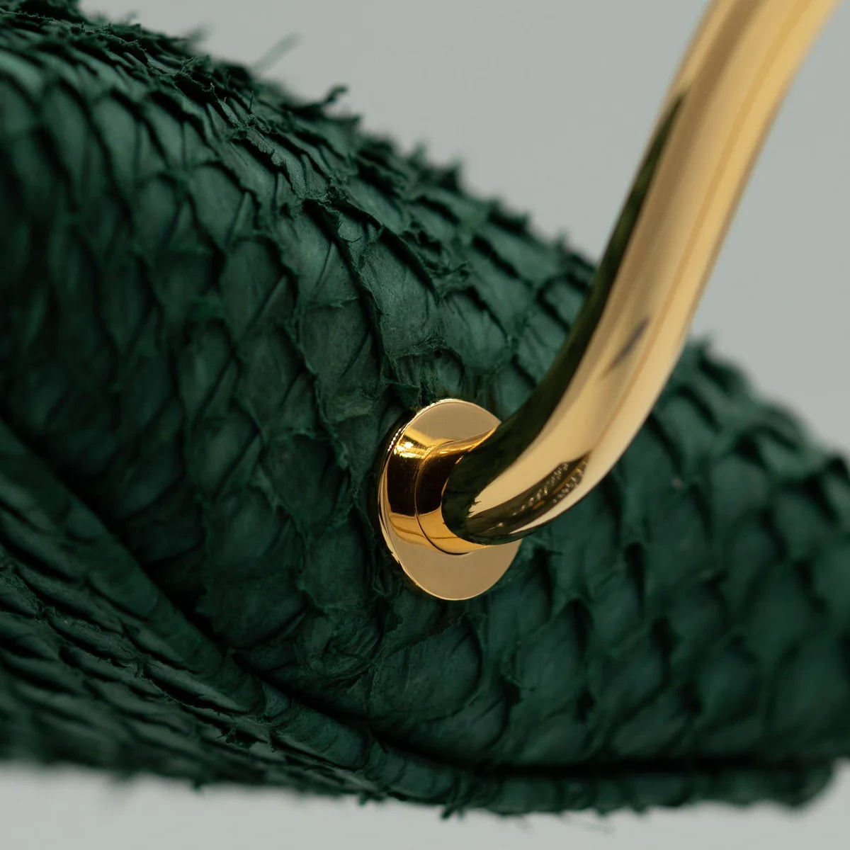 A close-up view of a green textured material with a shiny gold metal handle attached to it. The material appears to be woven or layered, and the handle is secured with a gold screw. The image highlights the contrast between the luxurious gold handle and the richly textured green surface.