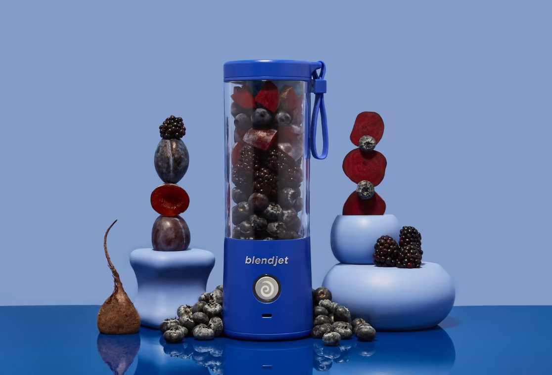 A deep blue BlendJet blender filled with blueberries, blackberries, and beets, surrounded by stacked plums, dried beet slices, and scattered berries, set against a monochromatic blue background.