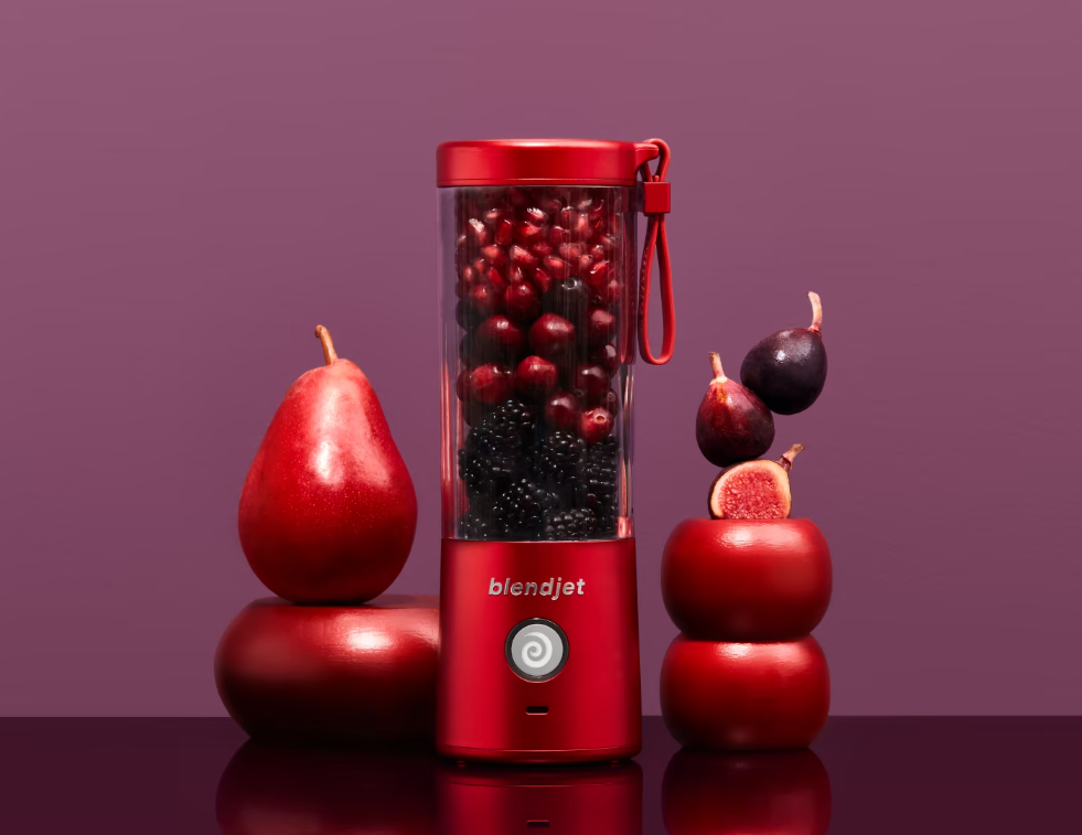 A bold red BlendJet blender filled with deep red fruits like cherries, pomegranate seeds, and blackberries, surrounded by fresh produce, including a glossy red pear, stacked red plums, and figs, set against a rich plum-colored background.