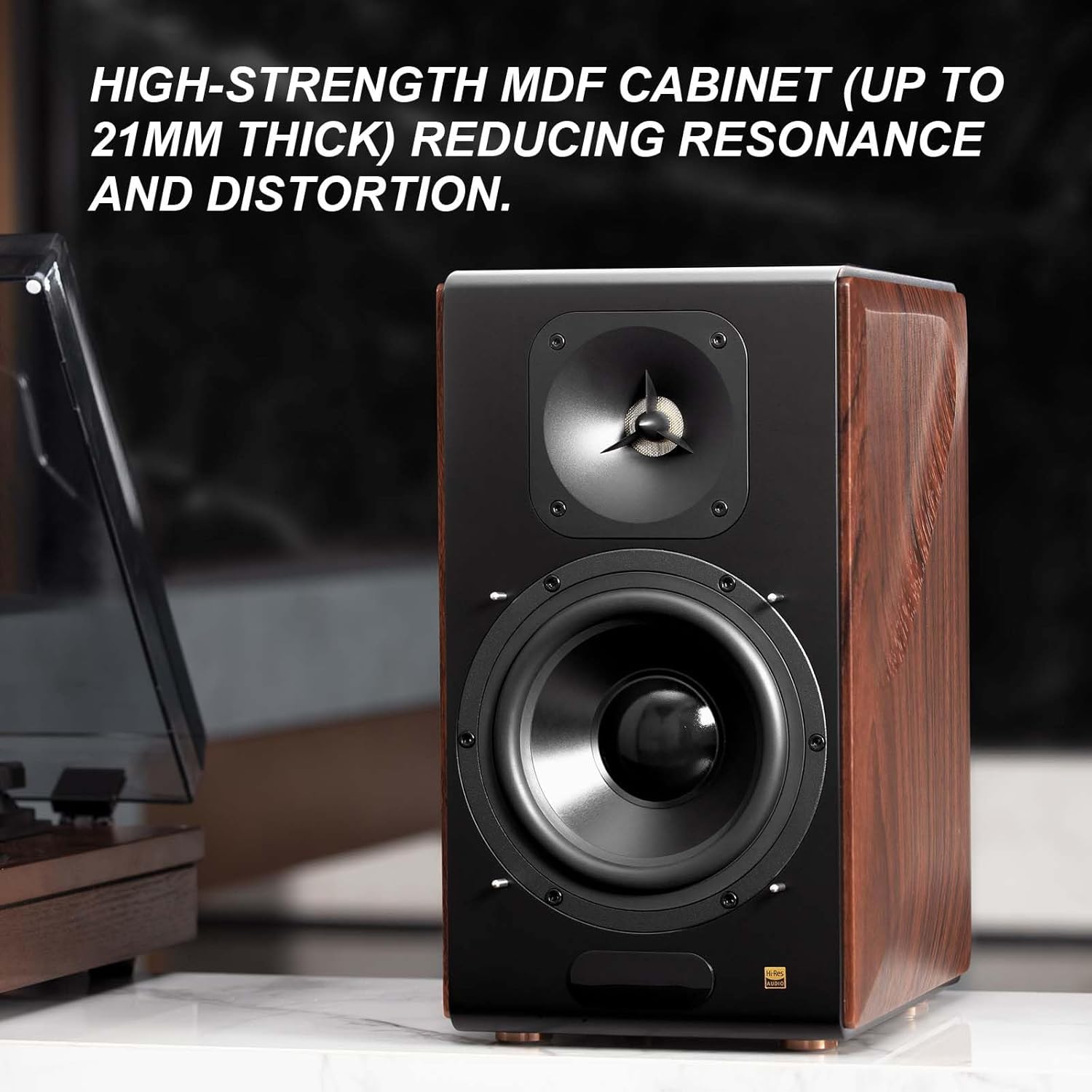 A premium bookshelf speaker with a high-strength MDF cabinet featuring a wood grain finish, designed to reduce resonance and distortion. The speaker has a sleek black front panel with a tweeter and woofer, emphasizing high-quality audio performance. The text highlights its 21mm thick cabinet construction, ensuring superior sound clarity and minimal vibration. A turntable is partially visible in the background, suggesting a high-fidelity audio setup.