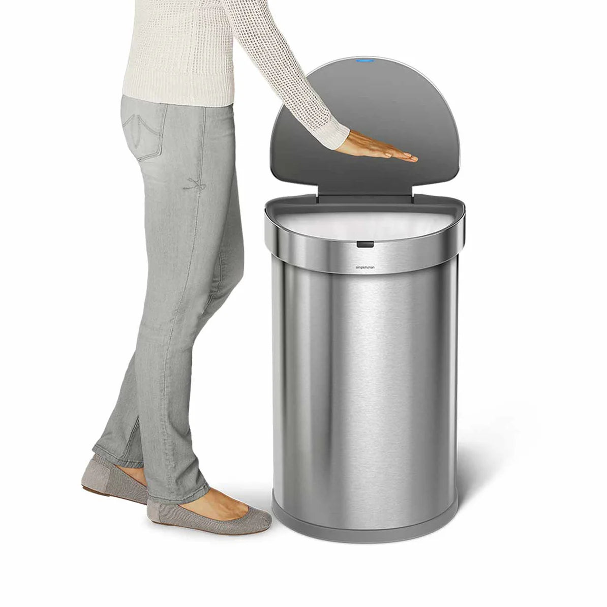 The image shows a Simplehuman touch-free sensor trash can made of stainless steel. A person is shown standing next to the trash can, with their hand hovering over the open lid, demonstrating the touch-free feature. The lid opens automatically when the hand is detected, which is a key feature of this product. The design is sleek and modern, with a cylindrical shape and a polished metal finish. The trash can is designed to be both functional and aesthetically pleasing, fitting well into a contemporary kitchen