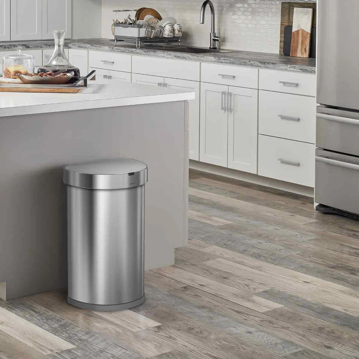 This image shows a modern kitchen setting where the stainless steel semi-round trash can is positioned next to a kitchen island. The trash can seamlessly integrates with the kitchen's contemporary design, which features light-colored cabinetry, a sleek countertop, and stainless steel appliances. The trash can's polished finish complements the overall aesthetic of the space, emphasizing both functionality and style. The placement near the island demonstrates its convenience for use in a busy kitchen environm