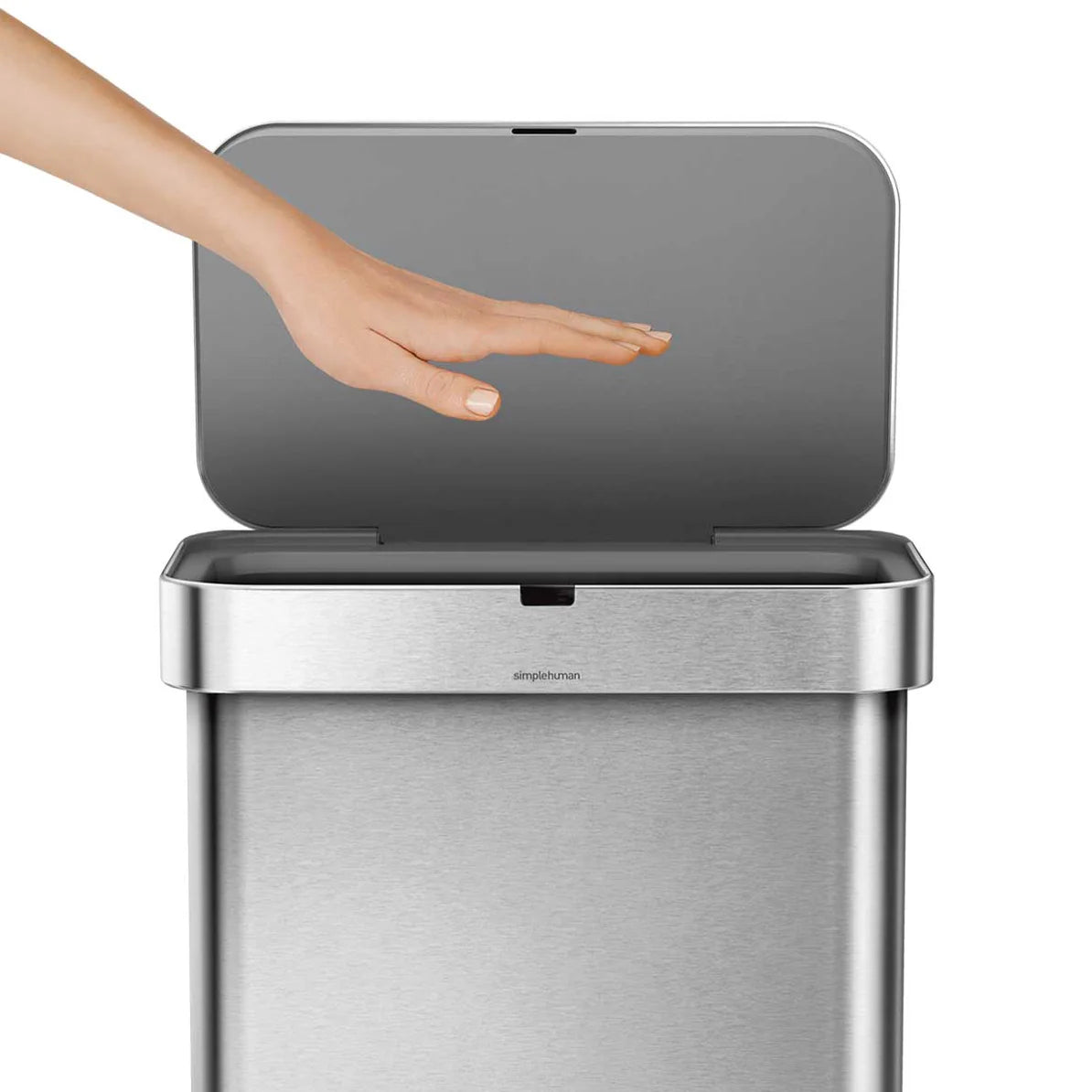 The image features a Simplehuman sensor trash can, which automatically opens its lid when a hand is waved over it. The can has a sleek, modern design with a brushed stainless steel finish. The hand is positioned above the sensor area, demonstrating the hands-free operation of the trash can. The lid is partially open, highlighting the responsive nature of the sensor technology, which activates without any physical contact. The Simplehuman branding is visible on the front of the trash can, reinforcing the pro