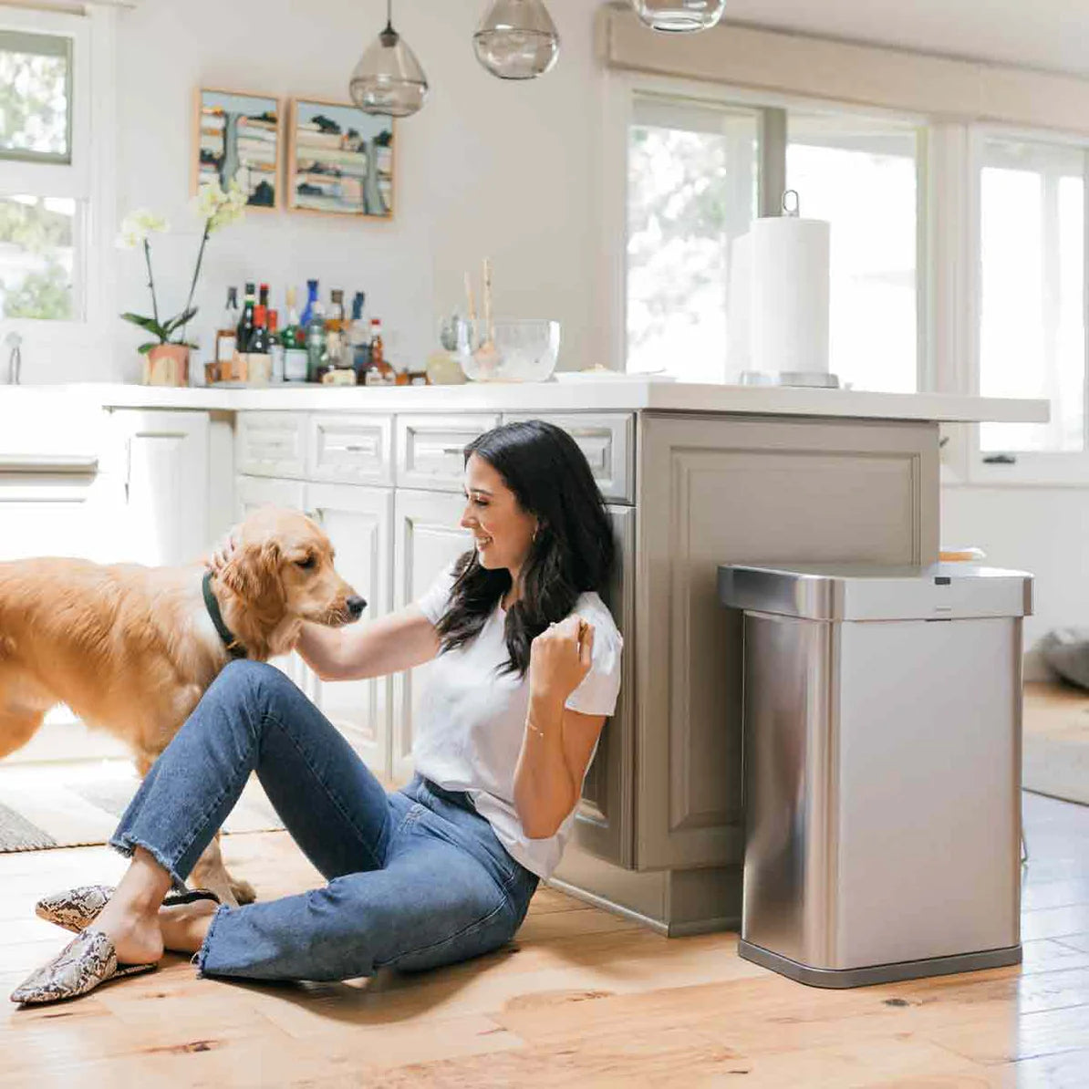 The image shows a cozy kitchen scene with a woman sitting on the floor, interacting with a golden retriever. The kitchen is bright and modern, featuring a large island in the center with cabinetry in a soft, neutral color. On the right side of the image, next to the island, is a Simplehuman trash can with a stainless steel finish. The trash can blends seamlessly into the kitchen’s modern design, adding a touch of elegance to the space.