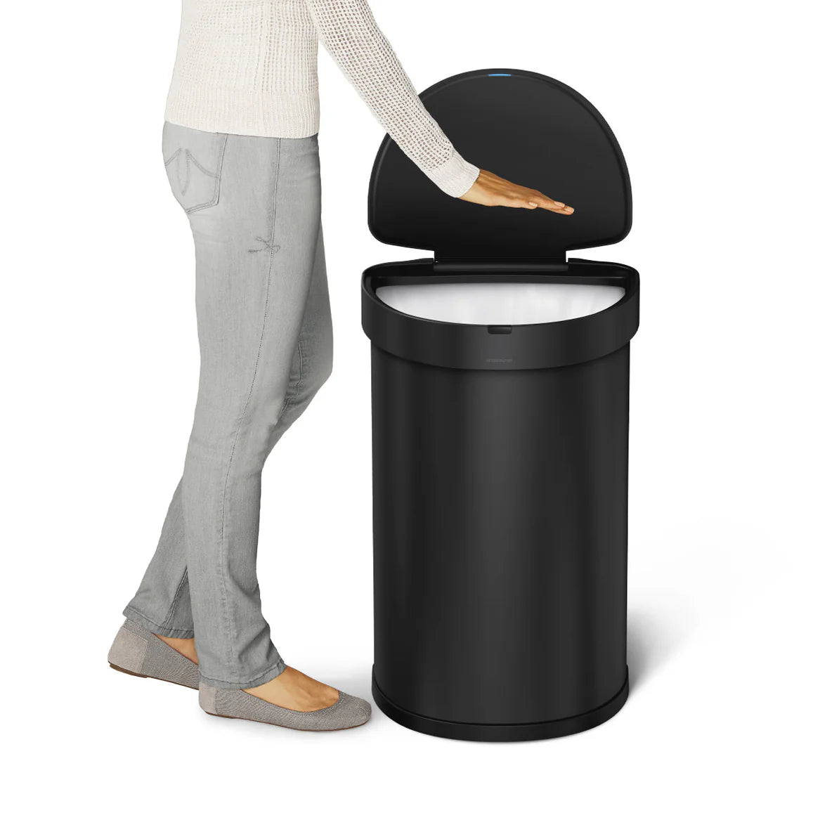 The image shows a Simplehuman trash can with a round shape and a sensor-activated lid. The trash can is black with a smooth, matte finish. The lid is slightly curved and opens automatically when a hand is placed above the sensor, demonstrating its touchless operation. A person standing next to the trash can, with their hand hovering over the sensor, is shown to illustrate the height and functionality of the trash can. The overall design is sleek and modern, suitable for various interior settings.