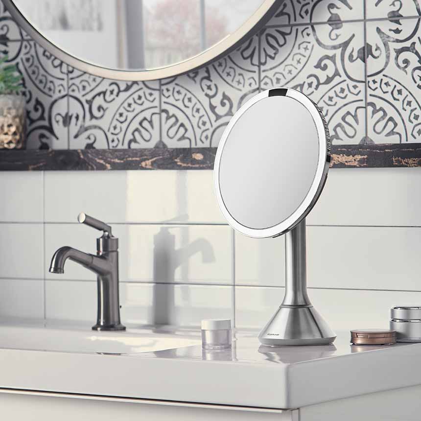 This image features a **Simplehuman makeup mirror** placed on a bathroom vanity. The mirror has a sleek, stainless steel finish, which complements the modern and sophisticated decor of the bathroom. The LED light ring around the mirror provides bright, even illumination, perfect for makeup application or grooming tasks.  The mirror is set against a backdrop of decorative tiles with a stylish, intricate pattern, adding to the overall aesthetic of the space. The vanity also includes other elements like a fauc