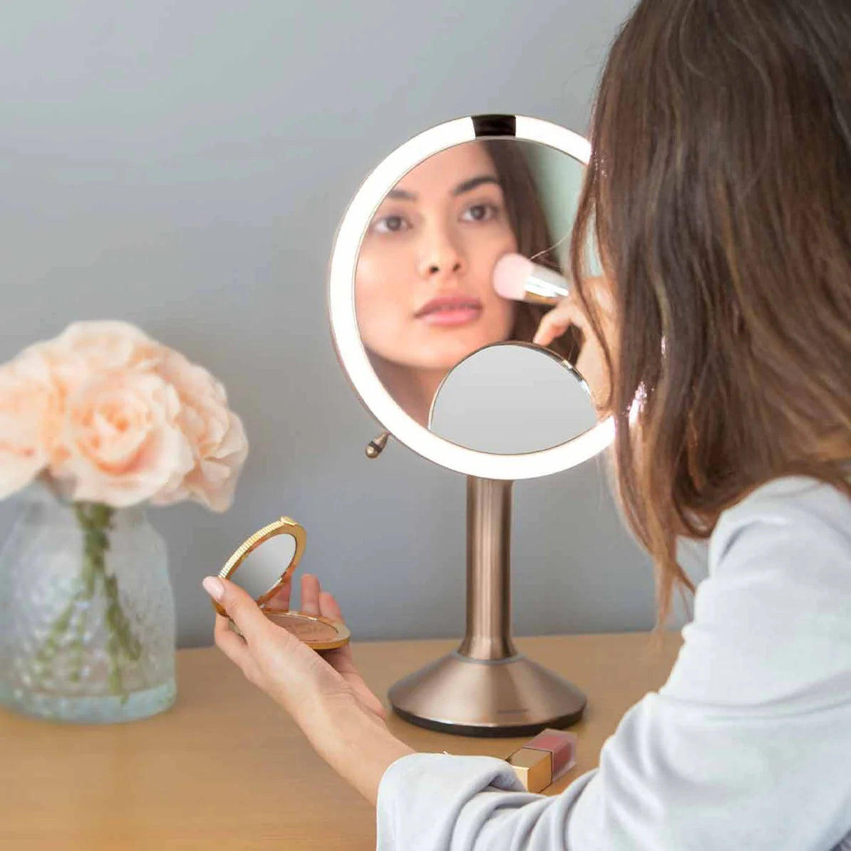 This image shows a woman using the Simplehuman makeup mirror as she applies makeup. The mirror features a bright LED light ring, providing clear, even illumination that enhances visibility. The reflection in the mirror highlights the mirror's ability to deliver true-to-life color and detail, which is essential for precise makeup application.  The mirror is placed on a vanity, where the woman is using it to blend makeup with a brush while holding a compact in her other hand. The design of the mirror stand is