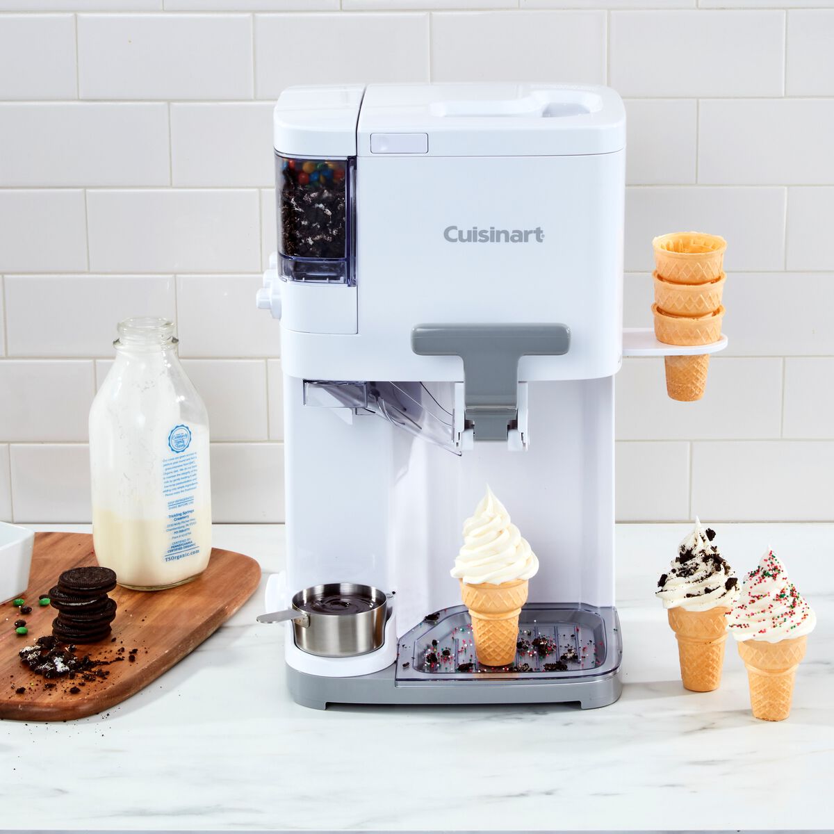 Cuisinart soft serve ice cream maker dispensing vanilla ice cream into a waffle cone, with Oreo toppings and milk nearby for preparing various flavors.