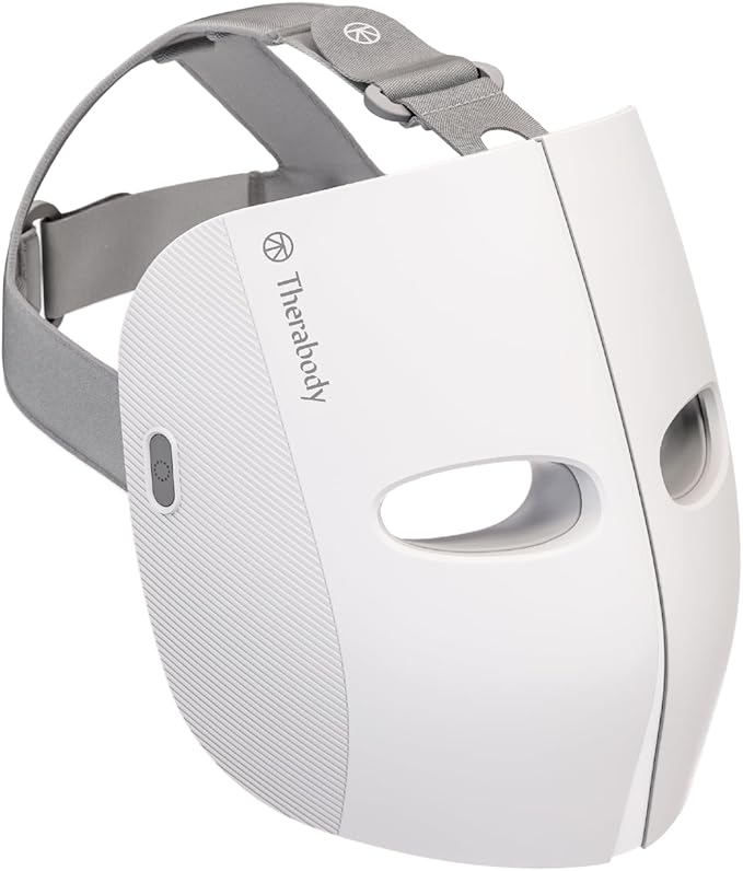 A close-up image of the Therabody TheraFace Mask, featuring a sleek white design with textured side panels and eye cutouts. The mask includes an adjustable gray head strap for a secure and comfortable fit. A power button is located on the side for easy operation. Designed for skincare treatments, the mask offers features such as red light therapy and vibration therapy to improve skin health and appearance.