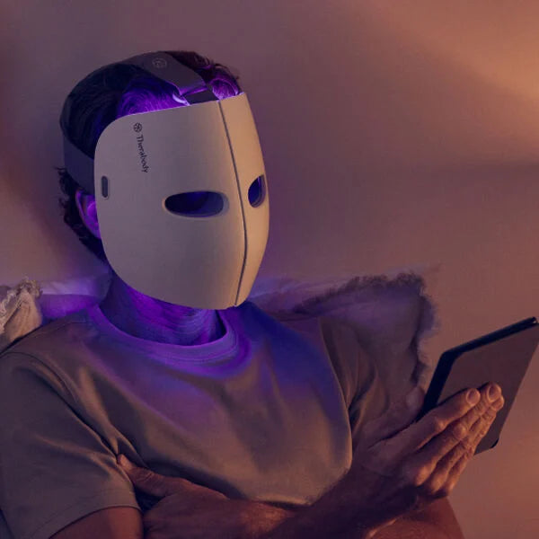 A person wearing a white LED therapy face mask while relaxing on a couch and reading a tablet. The mask emits a soft purple light and is secured with an adjustable head strap for a comfortable fit. The ambient lighting creates a calming atmosphere, highlighting the mask's use for skincare treatments aimed at improving skin health and appearance.