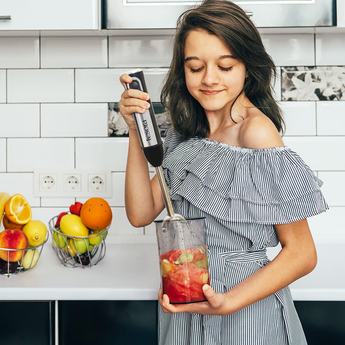 This image shows a young girl using a **hand blender** to blend fruits in a kitchen setting. The blender appears to be from the brand **Tesora**, making it a practical tool for preparing smoothies or purees. The kitchen background is bright and clean, with fruit baskets adding a vibrant touch. The scene portrays the blender as an easy-to-use and convenient appliance for both kids and adults in their everyday kitchen tasks.