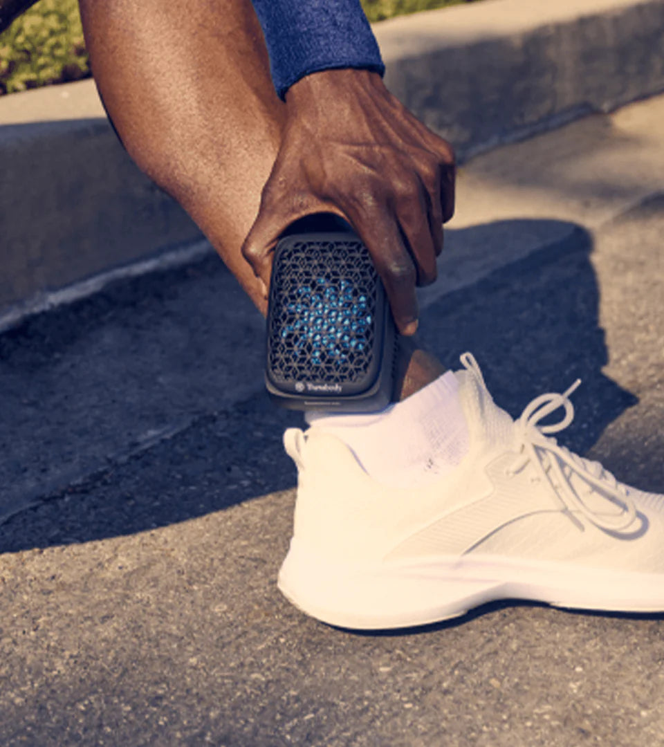 A person using a Therabody muscle therapy device with a hexagonal mesh design on their ankle outdoors, demonstrating its portability and effectiveness for targeted muscle recovery and pain relief.