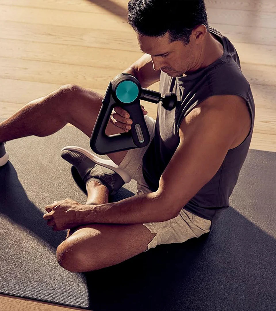 A man using a Theragun massage device on his leg while seated on a mat, showcasing its ergonomic design and effectiveness for deep muscle therapy and recovery.