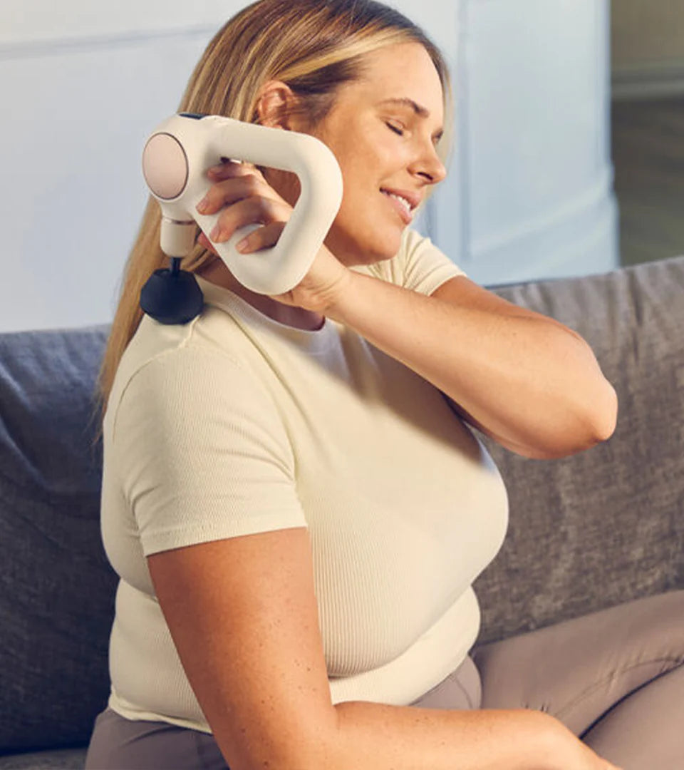 A woman using a handheld massage device on her shoulder while sitting on a couch, demonstrating its ergonomic design and ease of use for personal muscle relaxation.