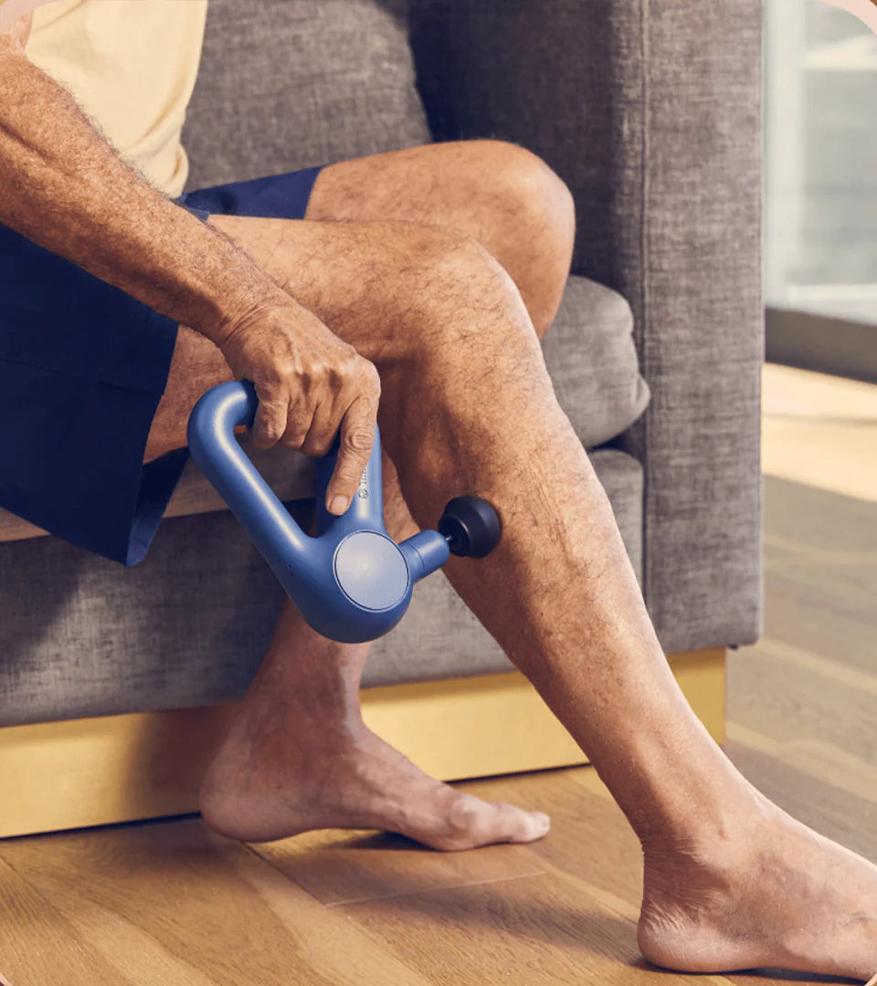 A person using a blue Theragun massage device on their calf while sitting on a couch, showcasing its ergonomic handle and effectiveness for muscle recovery and relaxation.