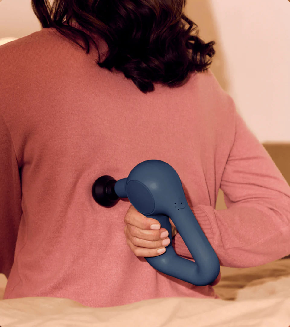 A woman using a blue Theragun massage device on her back, demonstrating its ergonomic design and ability to provide effective muscle relief in hard-to-reach areas.