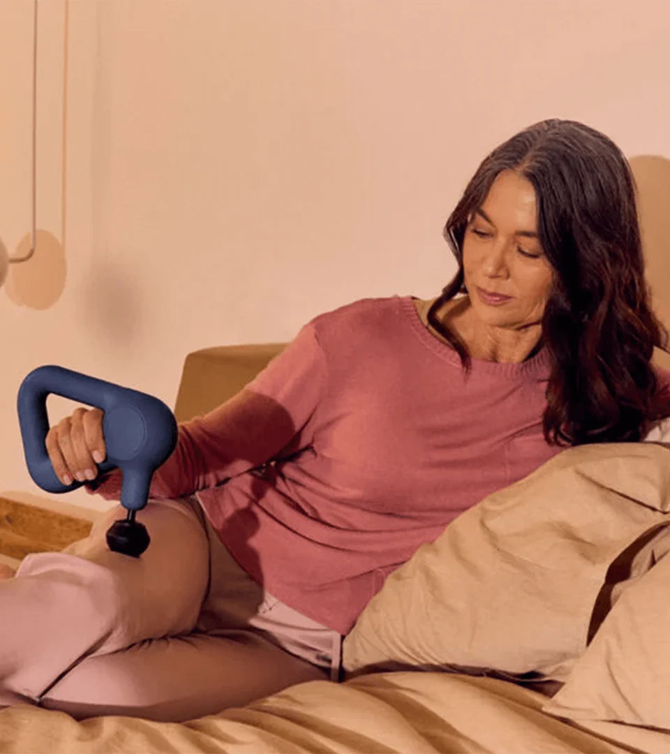 A woman using a blue Theragun massage device on her leg while sitting comfortably on a couch, demonstrating its ergonomic design and effectiveness for muscle relaxation and recovery.