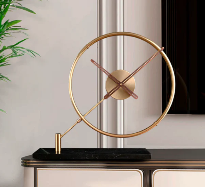 Elegant gold clock with a minimalist circular frame and slender hands, mounted on a sleek black base. The clock is displayed on a stylish console table against a soft neutral wall with panel detailing, accompanied by a hint of greenery in the background.