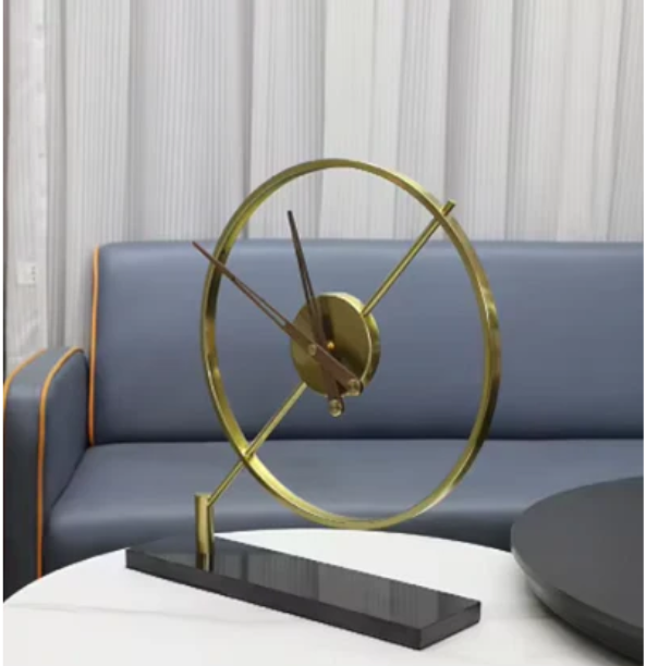 Modern gold-framed clock with sleek hands, set on a polished black base, displayed on a round white table. The backdrop features a gray sofa with orange piping and vertical pleated drapes, creating a chic and elegant ambiance.