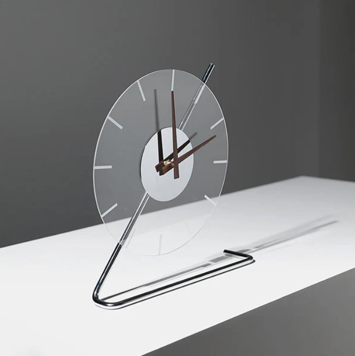 Contemporary clock with a clear circular face, minimal hour markers, and sleek hands, supported by a chrome metal stand, displayed on a simple white surface against a gray background for a clean, modern look.