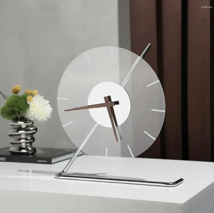 Modern glass clock with minimalist hour markers and elegant wooden hands, supported by a polished chrome stand, placed on a white table. The setup includes a silver decorative object and a floral arrangement in the background, enhancing the sleek and contemporary aesthetic.