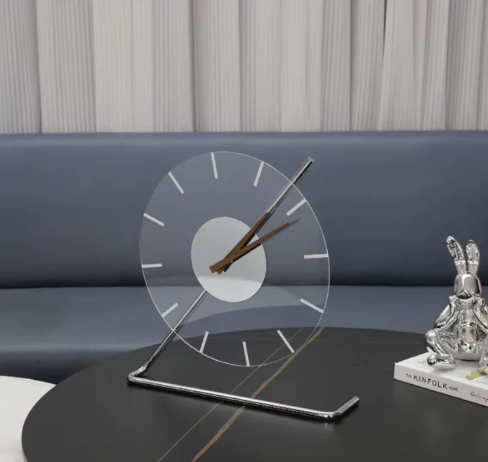 Modern minimalist clock with a transparent circular face and sleek metal stand, placed on a black tabletop next to a silver decorative figure and a book titled "The Kinfolk Home," set against a gray-blue sofa and neutral drapes.