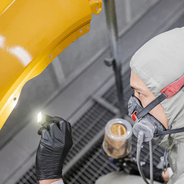  A person wearing a protective suit, gloves, and a respirator mask is using an LED clip light to inspect a large yellow-painted metal object. The setting appears to be an industrial or workshop environment, possibly related to manufacturing or maintenance. The LED light provides bright, focused illumination for detailed inspection.