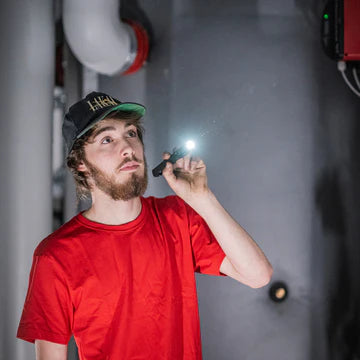 A man wearing a red shirt and a cap is holding a lit black LED penlight, illuminating a dark space. The background shows industrial pipes and equipment, indicating he is likely inspecting or working in an industrial environment. The light from the penlight is focused, providing a bright spot in the dark area.