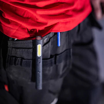  A close-up view of a man wearing a red shirt and black work pants, with a black LED penlight clipped to his pocket. The penlight features a long, narrow LED strip and a power button. Another pen is visible in the same pocket, indicating the practical use of carrying tools and accessories for work.