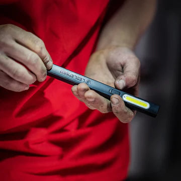  A close-up view of a person wearing a red shirt and holding a black LED penlight with a long, narrow LED strip. The person's hands are slightly dirty, suggesting they are engaged in manual or technical work. The penlight displays the brand "LED LENSER" on its body.