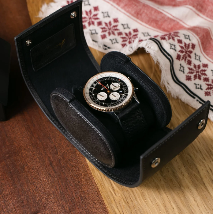 Open black leather watch case holding a sophisticated wristwatch with a black dial, white subdials, and a textured strap, set on a wooden surface next to a patterned fabric cloth.