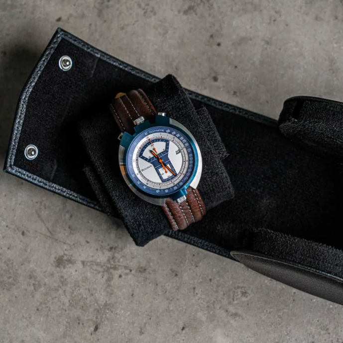 Open black leather watch case with a stylish wristwatch inside, featuring a white and blue dial, orange accents, and a brown leather strap, placed on a textured concrete surface.