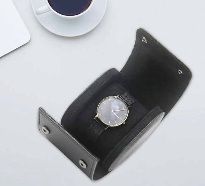 Black leather watch travel case opened to reveal a sleek wristwatch with a black mesh strap and gold accents, set on a soft interior, placed on a light surface beside a cup of coffee.