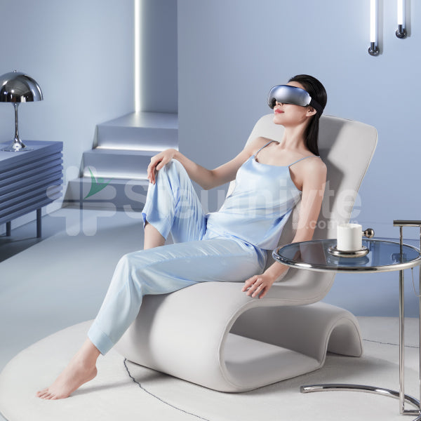 A woman wearing a reflective visor neck massager is relaxing in a modern, minimalist room. She is seated on a stylish white lounge chair, dressed in a light blue satin outfit, with her legs crossed and one hand resting on a nearby round side table holding a cup. The room features a sleek design with a metallic table lamp and ambient lighting on the walls.