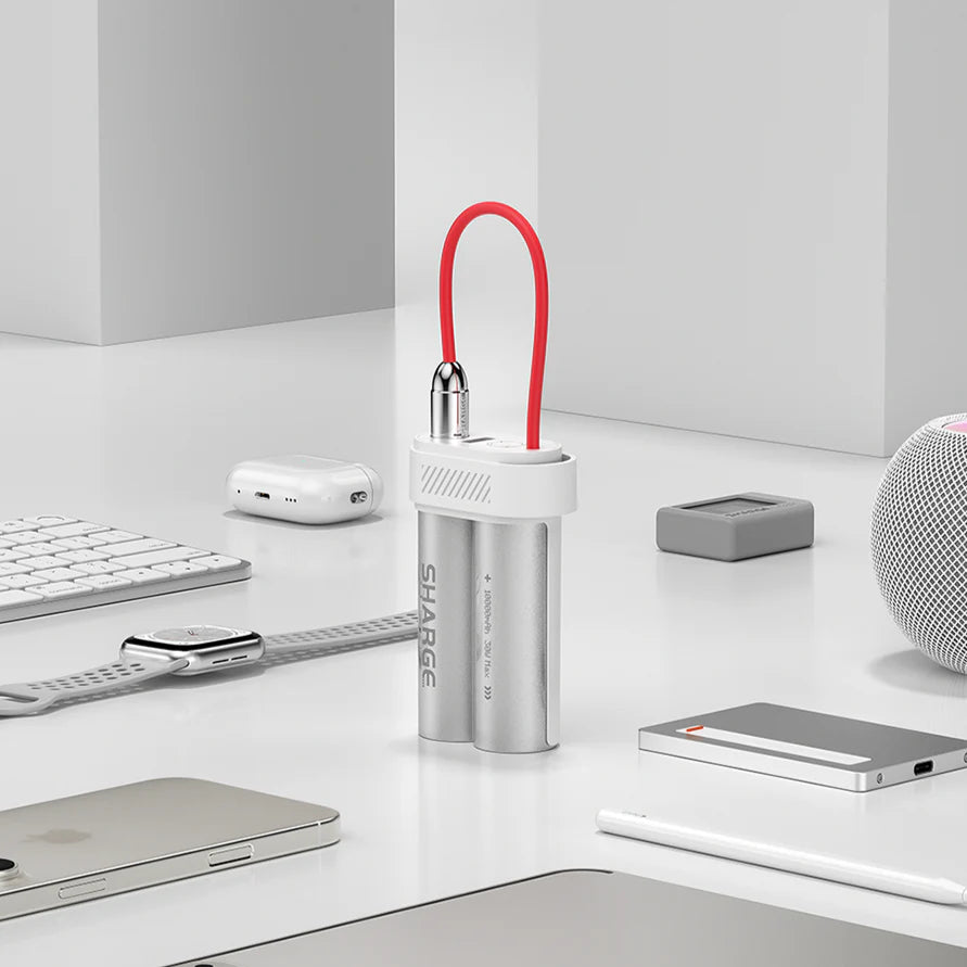 A SHARGE portable power bank styled like dual cylindrical batteries with a metallic silver finish, featuring a compact design and a bright red flexible charging cable. The power bank is placed on a clean, minimalistic workspace surrounded by tech accessories, including a smartwatch, keyboard, earbuds, and other devices. It blends seamlessly with modern gadgets, emphasizing its utility and stylish appeal for on-the-go charging.