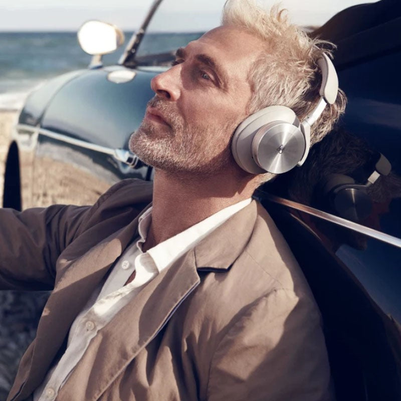 This image features a man relaxing outdoors, wearing **Bang & Olufsen over-ear headphones** in a **silver or light gray finish**. He is leaning against a classic car, capturing a serene moment with the ocean in the background. The scene emphasizes the **luxurious and high-end** nature of the headphones, with a focus on comfort, design, and **premium audio experience**. The natural setting, along with the man's casual yet stylish attire, enhances the idea of **effortless sophistication and lifestyle appeal**